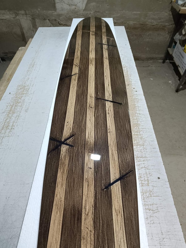 May include: A longboard deck featuring a striped pattern of alternating dark brown and light tan wood grain panels. The surface has a glossy, reflective finish and includes several dark, thin, cross-shaped inlay details embedded across the length.