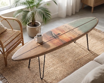 May include: A surfboard coffee table with black metal hairpin legs. The wooden surface features a sunset graphic, a silhouette of a surfer, and the text Cocoa Beach. A white ceramic mug containing a steaming hot beverage sits on the table.