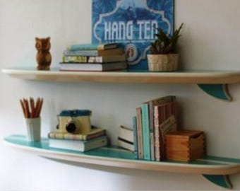 Op de afbeelding: Twee surfplankvormige planken aan een witte muur. De planken zijn van licht hout met een turquoise streep en vin. De bovenste plank bevat een klein uilbeeldje, boeken en een plant in een pot. De onderste plank heeft boeken, een vintage camera en potloden.