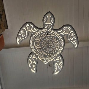 Może przedstawiać: Szara, podświetlana lampa w kształcie żółwia morskiego. Lampa posiada skomplikowane wycięcia i centralny motyw twarzy. Światło prześwituje przez wycięcia, tworząc efekt dekoracyjny. Lampa wisi na białym suficie.