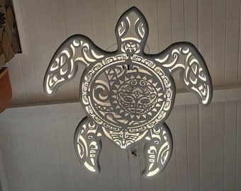 May include: A gray, illuminated sea turtle-shaped light fixture. The fixture features intricate cut-out designs and a central face motif. The light shines through the cut-outs, creating a decorative effect. The fixture is hanging from a white ceiling.