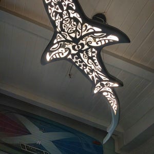 Puede incluir: Una lámpara de techo con forma de orca en blanco y negro con intrincados diseños recortados. La luz está encendida, mostrando los patrones detallados. Una tabla de surf es visible en el fondo.