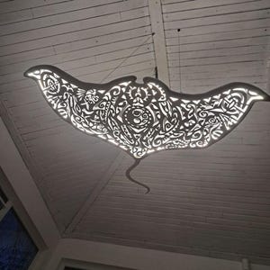May include: A decorative, manta ray-shaped light fixture. The fixture is suspended from the ceiling and features intricate cut-out patterns that allow light to shine through. The design is primarily white, with a curved tail and wing-like extensions.
