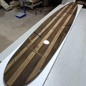 May include: A long, narrow surfboard featuring a striped pattern of alternating dark brown and light tan wood grain panels. The surface has a glossy, reflective finish with visible dark cross-shaped inlay details spaced along the length of the board.
