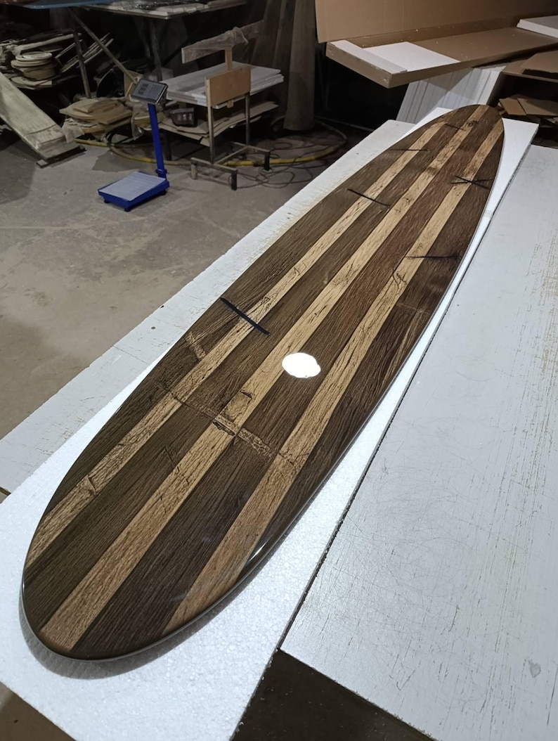 May include: A long, narrow surfboard featuring a striped pattern of alternating dark brown and light tan wood grain panels. The surface has a glossy, reflective finish with visible dark cross-shaped inlay details spaced along the length of the board.