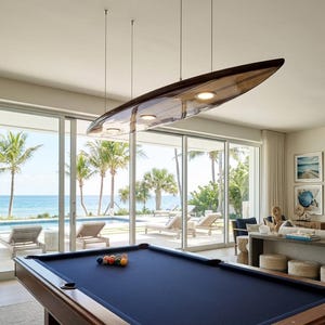 May include: A dark wood pool table with a navy blue felt surface sits beneath a custom pendant light fixture shaped like a surfboard. The light fixture is suspended by thin metal cables and features three integrated circular light bulbs.