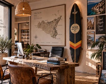 May include: A rustic wooden desk with a computer, books, and chairs in an office space. A large map of Sicily hangs on the wall next to a dark surfboard with yellow and red stripes. Framed travel photos and text signs decorate the walls.
