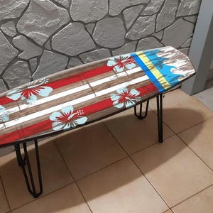 May include: A surfboard-shaped bench with a wooden top featuring red and white stripes, blue and white hibiscus flowers, and a blue and yellow wave design. The bench has black metal hairpin legs.