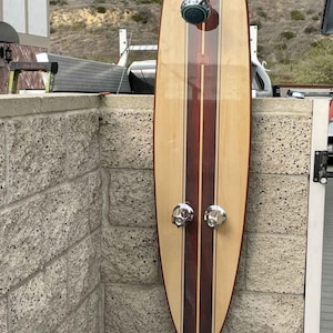May include: An outdoor shower with a surfboard design made of wood. The shower has a light wood grain with two dark brown stripes. It features a chrome shower head and two chrome knobs. The shower is mounted on a brick wall.
