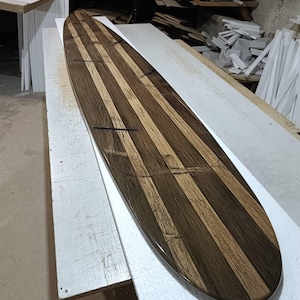 May include: A long, narrow surfboard constructed from alternating vertical strips of dark brown and light tan wood. The board features a smooth, polished finish with visible wood grain patterns and thin, dark horizontal inlay lines across the surface.
