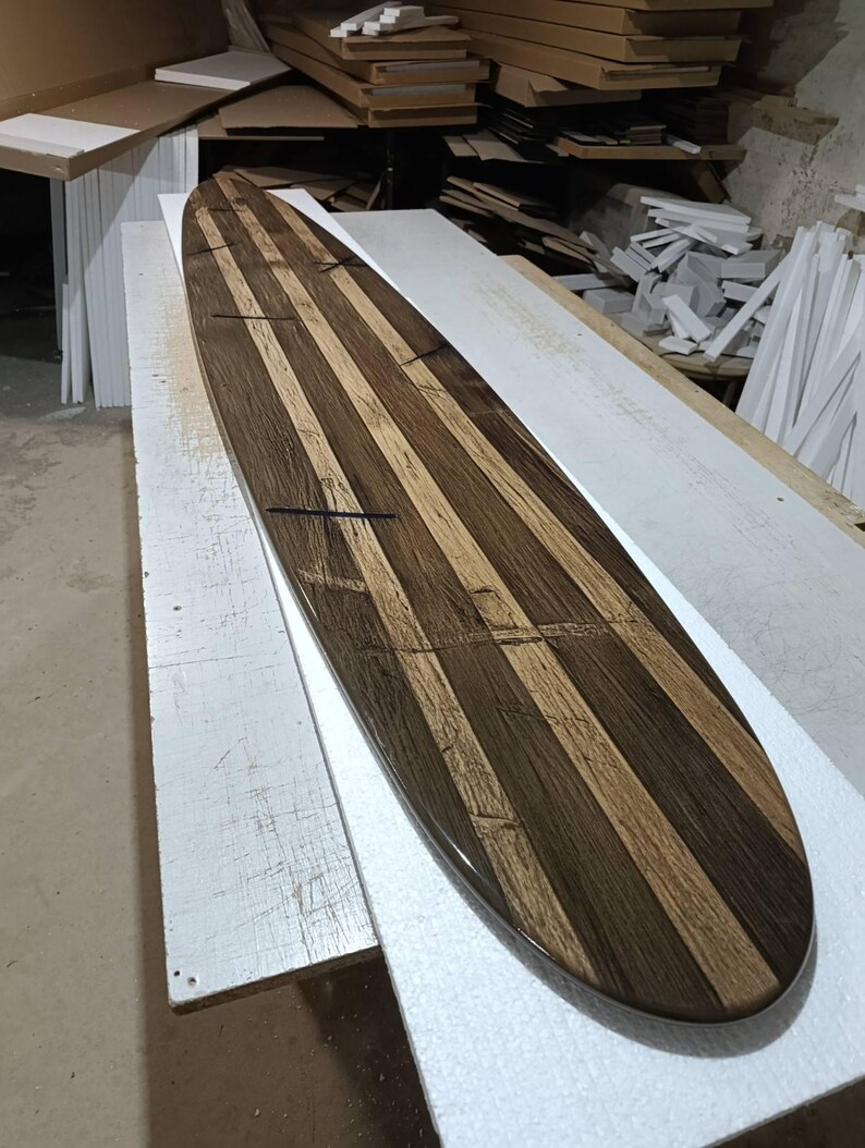 May include: A long, narrow surfboard constructed from alternating vertical strips of dark brown and light tan wood. The board features a smooth, polished finish with visible wood grain patterns and thin, dark horizontal inlay lines across the surface.