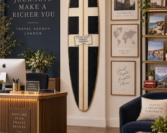 May include: A tall, vertical surfboard wall decoration featuring a black and white cross pattern with a central plaque reading FISTRAL BEACH NEWQUAY CORNWALL 1964. The surfboard is mounted on a white wall next to framed travel-themed wall art.