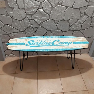 Puede incluir: Mesa de centro ovalada con diseño de tabla de surf. La tapa es blanca y azul con las palabras "Surfing Camp" y "Carolina Beach, NC" impresas. La mesa tiene patas de horquilla negras.