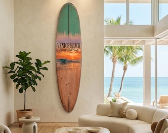 May include: A vertical surfboard wall art piece featuring a gradient design that transitions from a teal top to a sunset ocean scene in the middle and a natural wood grain finish at the bottom. The text Venice Beach Florida Gulf Coast is printed.