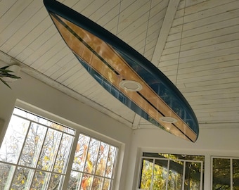 May include: A surfboard suspended from a white ceiling. The board has a blue top edge, a light brown center, and black stripes. Two circular lights are visible on the board's underside. Large windows with white frames and a view of trees are in the background.