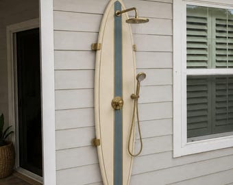 May include: An outdoor shower system mounted on a surfboard-shaped panel. The surfboard is cream-colored with a central blue stripe. It features a brass-finished shower head, a handheld shower wand, and a single control handle in brass.