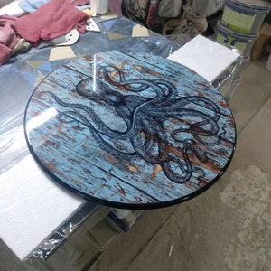 Op de afbeelding: Ronde houten tafelblad met een blauw en bruin octopus ontwerp. Het tafelblad heeft een zwarte rand.