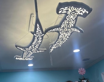 May include: Two hammerhead shark-shaped pendant lights suspended from the ceiling. Each light fixture features a dark grey exterior with intricate, cut-out tribal-inspired patterns that allow bright white light to shine through the body.