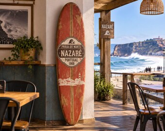May include: A tall, weathered red surfboard leaning against a wall, featuring a central emblem with the text PRAIA DO NORTE NAZARÉ PORTUGAL 1968, a graphic of a lighthouse, and a wave icon, displayed in a coastal setting overlooking the ocean.