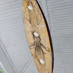 May include: A surfboard with a squid illustration. The board has a light brown wood grain design with a dark brown edge. Three white circular holes are visible on the board. The squid illustration is in black and brown.