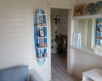 May include: A surfboard-shaped wall shelf with blue and white stripes and the letters "S", "B", and "R". The shelf has multiple levels holding books, a framed photo, and decorative items. The shelf is mounted on a white wall.
