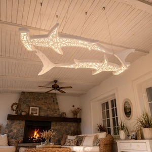 Può includere: Due lampade a sospensione bianche a forma di squalo martello appese a un soffitto in legno bianco. Ogni apparecchio presenta intricati motivi in filigrana tagliati al laser che lasciano filtrare una luce calda, creando un effetto decorativo.