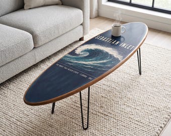 May include: A surfboard-shaped coffee table with a dark blue surface featuring a graphic of a crashing ocean wave. The text reads SEBASTIAN INLET and THE WAVE THAT NEVER STOPS - FLORIDA 1961. It has a wooden edge and four thin black metal hairpin legs.