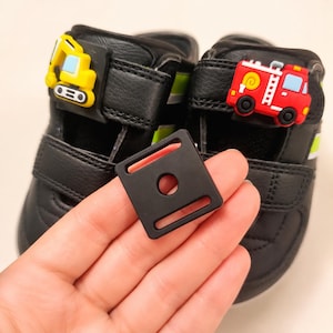 Könnte beinhalten: Ein Paar schwarze Sneaker mit einem gelben Bagger-Anhänger an einem Riemen und einem roten Feuerwehrauto-Anhänger am anderen. Eine quadratische schwarze Kunststoffschnalle mit einem Mittelloch und zwei rechteckigen Schlitzen liegt vorne.