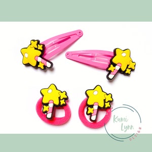 Könnte beinhalten: Vier Haaraccessoires: zwei rosa Haarspangen und zwei rosa Haargummis. Jedes hat einen gelben Stern mit einem Zuckerstangen-Design. Die Accessoires befinden sich auf weißem Hintergrund.