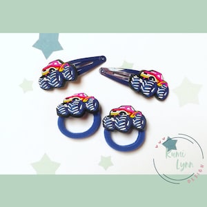 Könnte beinhalten: Vier blaue Haarschmuckstücke mit rosafarbenen und schwarzen Monstertruck-Designs. Zwei der Accessoires sind Haarklammern und zwei sind Haargummis.