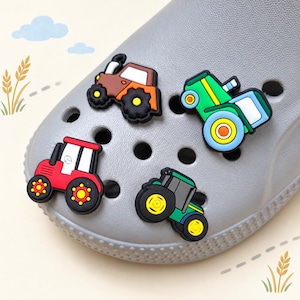 Könnte beinhalten: Vier traktorförmige Schuhanhänger an einem grauen, perforierten Clog. Die Anhänger zeigen einen braunen Traktor, einen grün-blauen Traktor, einen roten Traktor und einen dunkelgrünen Traktor, jeweils mit schwarzen Rädern und detaillierten Kabinen.