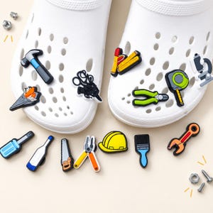 Könnte beinhalten: Ein Paar weiße Clogs, verziert mit verschiedenen bunten Schuh-Anhängern in Form von Bauwerkzeugen, darunter ein Hammer, eine Zange, ein Maßband, eine Säge, ein Schutzhelm, ein Pinsel, ein Schraubenschlüssel und ein Schraubendreher sowie lose Muttern.
