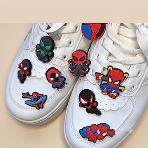 Könnte beinhalten: Ein Paar weiße Sneaker, verziert mit mehreren bunten Schuhanhängern aus Gummi, die verschiedene Spider-Man-Charakterdesigns in Rot, Blau, Schwarz und Grün zeigen. Die Anhänger sind an den weißen Schnürsenkeln und der Vorderseite befestigt.
