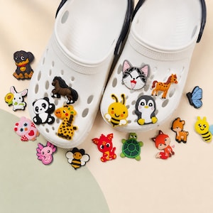 Könnte beinhalten: Ein Paar weiße Clogs, verziert mit verschiedenen bunten tierförmigen Charms, darunter ein Panda, eine Giraffe, ein Pferd, eine Katze, ein Pinguin, eine Biene, eine Schildkröte, ein Fuchs und ein Drache. Weitere lose Charms in Form von Hund, Hase, Eule, Axolotl und Schmetterling liegen verstreut.