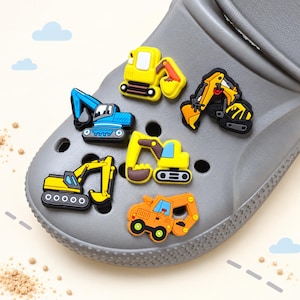 Könnte beinhalten: Ein grauer Clog-Schuh, verziert mit fünf bunten Gummi-Anhängern in Form von Baustellenfahrzeugen, darunter gelbe, blaue und orangefarbene Bagger und Traktoren, jeweils mit detaillierten mechanischen Teilen und Kettenlaufwerken.