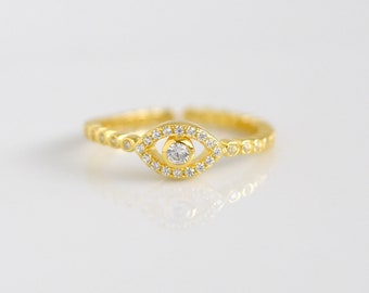 May include: A gold ring with a clear stone in the center, surrounded by smaller clear stones. The ring is in the shape of an eye.