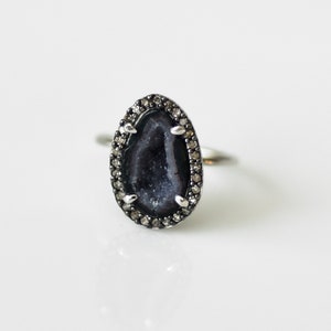 Puede incluir: Un anillo de plata con una piedra de ágata negra engastada en un halo de diamantes.