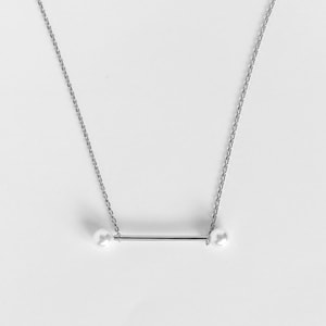 May include: A silver necklace featuring a horizontal bar with a pearl at each end. The chain is delicate and the pearls are white. The necklace is displayed against a white background.