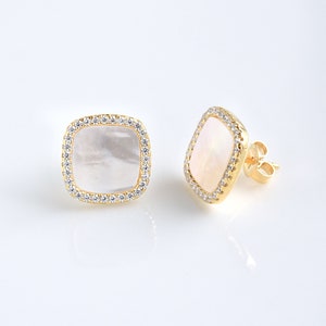 May include: A pair of gold stud earrings. Each earring has a square, iridescent, mother-of-pearl centre stone, encircled by a halo of small, clear crystals. The earrings are set on gold posts.