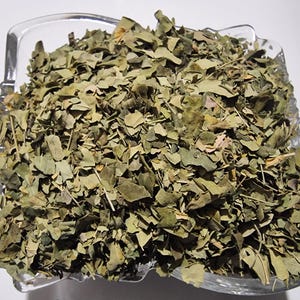 May include: A clear, square glass bowl filled with a large quantity of dried, crushed green leaves. The leaves are small, brittle, and vary in shades of olive and sage green, with some thin, light brown stems mixed throughout the pile.