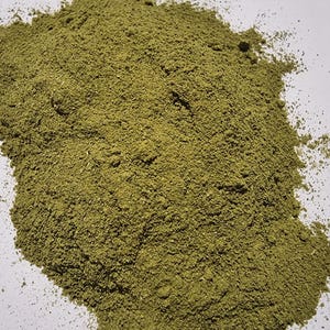 May include: A pile of finely ground, olive-green herbal powder with a dry, granular texture. The substance is spread in an irregular, organic shape, showing consistent coloration and a matte finish throughout the entire mound of loose botanical material.