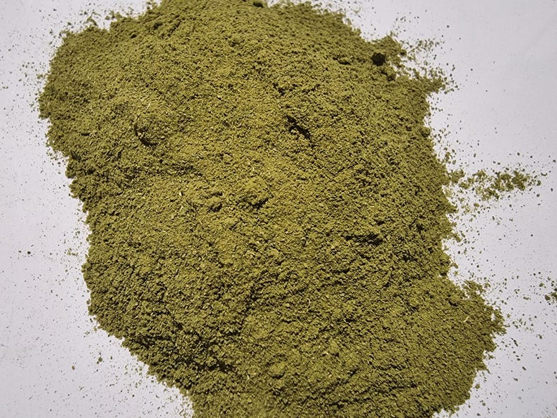 May include: A pile of finely ground, olive-green herbal powder with a dry, granular texture. The substance is spread in an irregular, organic shape, showing consistent coloration and a matte finish throughout the entire mound of loose botanical material.