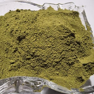 May include: A clear glass bowl filled with a fine, olive-green powder. The bowl has a square shape with rounded corners and a decorative, molded rim. The powder is piled high in the center, showing a textured, granular surface throughout the container.