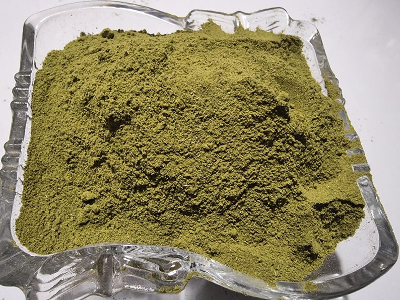 May include: A clear glass bowl filled with a fine, olive-green powder. The bowl has a square shape with rounded corners and a decorative, molded rim. The powder is piled high in the center, showing a textured, granular surface throughout the container.