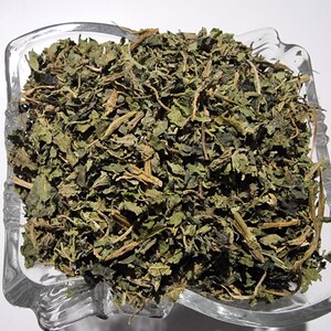 May include: A clear glass bowl filled with a heap of dried green herbs. The mixture consists of small, crinkled, dark green leaves and thin, light brown woody stems. The bowl has a square shape with rounded corners and a textured, decorative rim.
