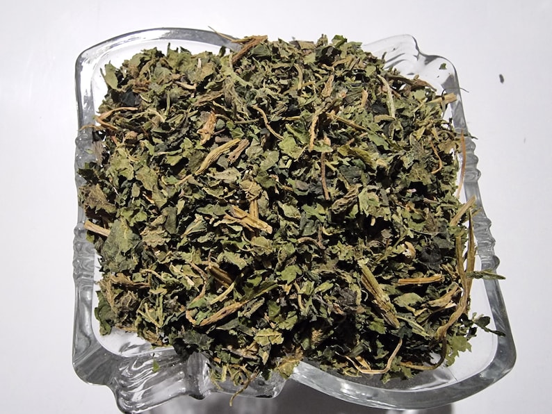May include: A clear glass bowl filled with a heap of dried green herbs. The mixture consists of small, crinkled, dark green leaves and thin, light brown woody stems. The bowl has a square shape with rounded corners and a textured, decorative rim.