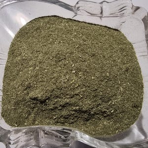May include: A clear, square glass bowl with scalloped edges filled with a finely ground, olive-green herbal powder. The texture of the powder appears dry and consists of small, uniform particles with occasional tiny, lighter-colored fibrous bits.