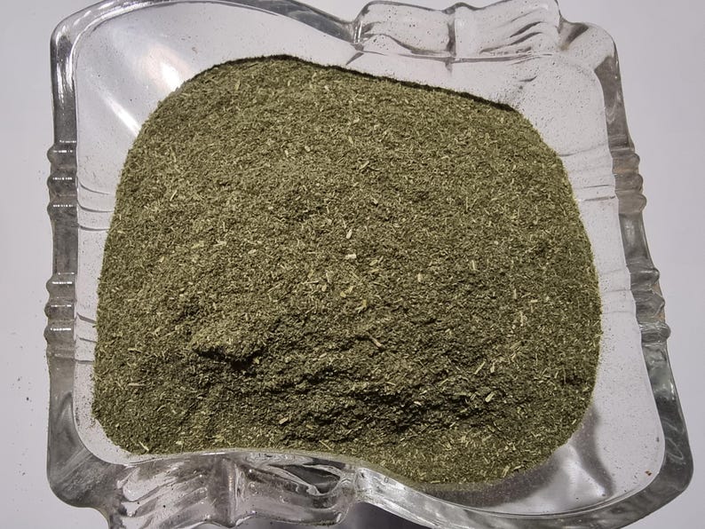 May include: A clear, square glass bowl with scalloped edges filled with a finely ground, olive-green herbal powder. The texture of the powder appears dry and consists of small, uniform particles with occasional tiny, lighter-colored fibrous bits.