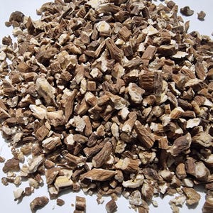 May include: A pile of dried, chopped root pieces featuring a mix of dark brown bark and light cream-colored interiors. The fragments are irregular in shape and size, scattered across a plain white surface, showing a fibrous and woody texture.