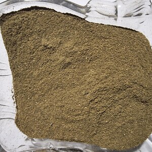 May include: A finely ground, earthy brown herbal powder contained within a clear, square glass dish with decorative scalloped edges. The texture of the powder appears uniform and dry, filling the center of the transparent glass container.
